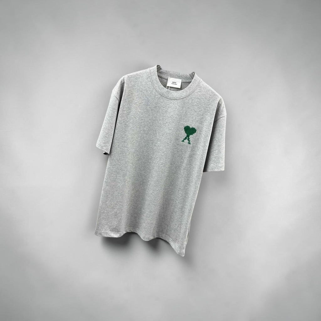 Model wearing an oversized grey AMI Paris t-shirt with a green heart logo at a Parisian cafe.