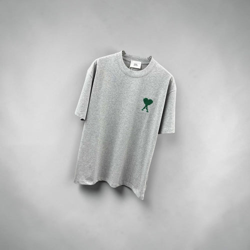 Model wearing an oversized grey AMI Paris t-shirt with a green heart logo at a Parisian cafe.