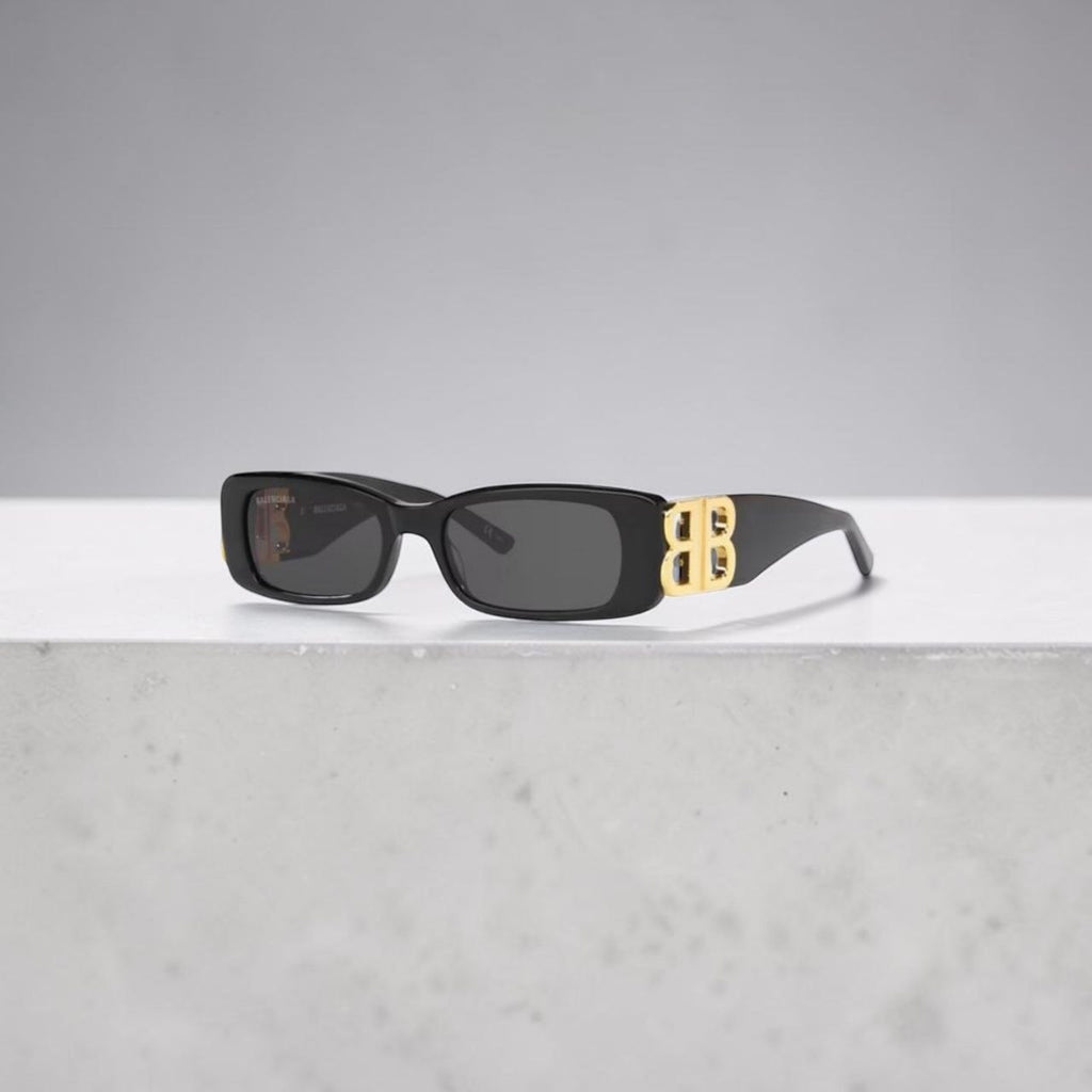 Model wearing black Balenciaga Dynasty rectangle sunglasses with gold BB logos on a city street.