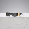 Model wearing black Balenciaga Dynasty rectangle sunglasses with gold BB logos on a city street.