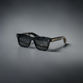 Model wearing thick black Jacques Marie Mage Torino rectangular sunglasses in a city setting.